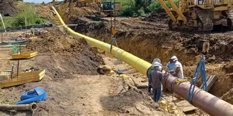 Long yellow-sleeved pipeline section positioned in an excavated trench with plant on standby